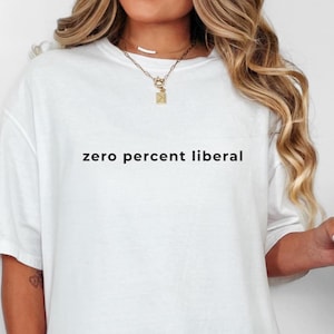 May include: White oversized t-shirt with the text "zero percent liberal" in black. The shirt is paired with a clear glass tumbler with a wooden lid and a straw. The person is wearing gold jewellery.