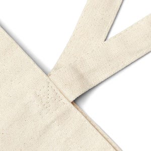 May include: Close-up of the top of a beige canvas tote bag with a reinforced seam at the top of the handle.