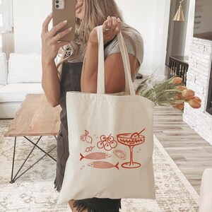 May include: A white canvas tote bag with a red illustration of a cocktail glass, two fish, an oyster, lemons, and cherries. The bag has two handles.