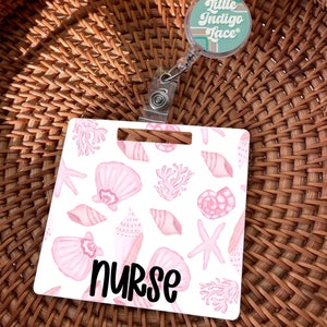 May include: A square badge reel featuring a white background with a repeating pattern of light pink seashells, starfish, and coral. The word nurse is printed in black script at the bottom. A circular reel above displays the text Little Indigo Lace.