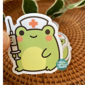 May include: A sticker featuring a cartoon frog wearing a nurse's cap with a red cross and holding a syringe. The frog is light green with a yellow belly and pink cheeks. The sticker has the text "little indigo lace".