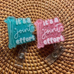 May include: Two acrylic badge reels featuring a glittery finish, one in teal and one in pink. Each badge reel displays a white illustration of a bone and the text "it's a joint effort" in a combination of block and script lettering with small star accents.