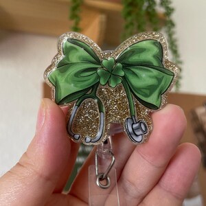 Puede incluir: Un carrete de insignia de brillo dorado con un trébol de cuatro hojas y un lazo verde. El carrete tiene un clip de plástico transparente y un anillo de metal.