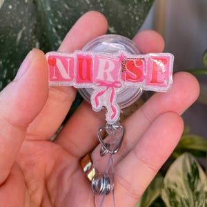 Peut inclure: Un porte-badge rose et blanc avec le mot "NURSE" dans un design en lettres majuscules. Un graphique de stéthoscope et de nœud rose est en dessous du mot. Le porte-badge a un cordon rétractable en plastique transparent et un clip en métal.
