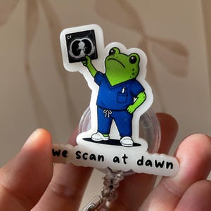 May include: A circular badge reel featuring a green frog wearing blue medical scrubs and white shoes. The frog holds a CT scan image above its head. Below the frog, a white speech bubble-shaped banner displays the black text we scan at dawn.