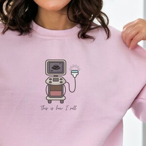 May include: Light pink sweatshirt featuring a medical ultrasound machine graphic. The illustration includes a screen, controls, and a probe, with the text "This is how I roll" below. The design is in shades of brown, gray, and white.