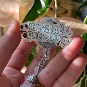 May include: A clear badge reel with the words "PHYSICAL THERAPY" in light blue script. The badge features illustrations of dumbbells, a kettlebell, and a walker. The reel has a clear plastic clip and a silver metal clasp.
