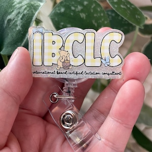 May include: A glittery acrylic badge reel featuring the text IBCLC in a yellow and white checkered pattern. The design includes a small bear illustration, a blue butterfly, and the text international board certified lactation consultant.