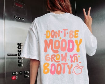 May include: White t-shirt with the text "DON'T BE MOODY GROW YA BOOTY" in orange, pink, and yellow. A peach graphic is below the text. The shirt has a relaxed fit.