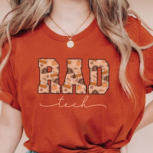 May include: Rust-colored t-shirt with the words "RAD tech" in a block letter design. The letters are filled with a fall-themed pattern of pumpkins, pie slices, and floral elements. The word "tech" is in a cursive font.
