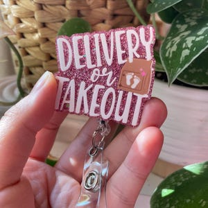 May include: A pink glitter badge reel featuring the white text DELIVERY or TAKEOUT and a brown paper bag graphic with white baby footprints. The badge reel has a clear plastic strap and a metal clip mechanism for attaching to clothing or accessories.