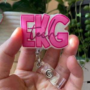 May include: A pink and white EKG tech badge reel. The badge reel features the words "EKG tech" in pink lettering. The badge reel is attached to a clear plastic retractable reel.