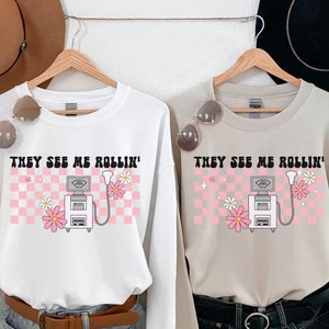 May include: Two long-sleeved sweatshirts, one white and one beige, with the text "THEY SEE ME ROLLIN'" above a graphic of an ultrasound machine, pink flowers, and a pink and white checkered background.