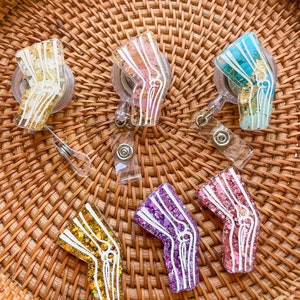Pu&ograve; includere: Sei portachiavi in resina colorati a forma di osso del ginocchio umano con glitter e scaglie dorate. I portachiavi sono su una superficie di vimini intrecciato.