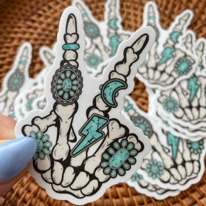 May include: A white sticker featuring a skeleton hand making a peace sign. The design includes turquoise accents, such as a lightning bolt, floral shapes, and crescent moons. The sticker is held against a woven basket background.