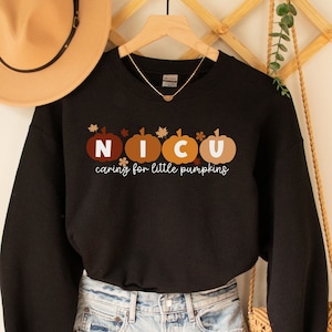May include: Black sweatshirt with the text "NICU caring for little pumpkins" in white and brown. The design features pumpkin illustrations in shades of brown and orange. The sweatshirt is hanging on a wooden hanger.