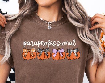 May include: Espresso-colored t-shirt with the word "paraprofessional" in white script above four orange pumpkins, each adorned with a different patterned bow. The bottom left corner of the shirt displays the word "ESPRESSO" in a circular design.