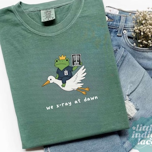 May include: A sage green t-shirt featuring a graphic of a frog wearing a yellow crown and blue scrubs, riding a white stork while holding an x-ray of a skeleton. The text below the graphic reads, we x-ray at dawn, in white lowercase letters.