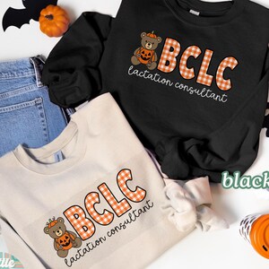 May include: Black and sand-colored sweatshirts with the text "BCLC lactation consultant" in orange gingham letters. A teddy bear wearing a pumpkin hat is next to the text. Halloween-themed apparel.