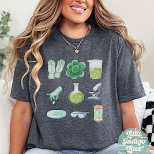 May include: Dark heather grey t-shirt with a science theme. The shirt features a variety of science-related illustrations, including a beaker, a petri dish, a microscope, and a pair of gloves. The text "Little Indigo Lace" is visible on a sticker in the lower right corner of the image.