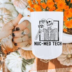 May include: White ceramic mug with a black and white illustration of a skeleton holding a cup with a radiation symbol. The text "NUC-MED TECH" is printed on the mug.