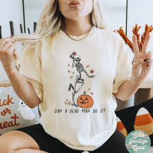 May include: A white t-shirt with a skeleton wearing a pumpkin hat and holding a leaf. The text "Can a dead man do it?" is printed below the skeleton. The shirt is on a model wearing black shorts and brown shoes.