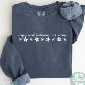 May include: A denim blue sweatshirt with the words "registered behavior technician" in white script, accented by a row of white daisies with yellow centers. The word "denim" is printed in a green font at the bottom left.