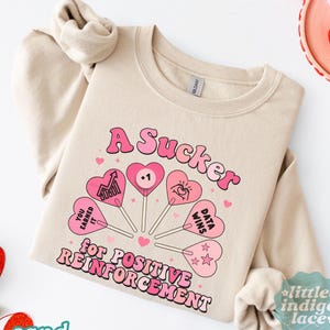 May include: Sand-colored sweatshirt with the text "A Sucker for Positive Reinforcement" in pink. The design includes heart-shaped lollipops with various graphics and text. The sweatshirt has long sleeves and a crew neck.