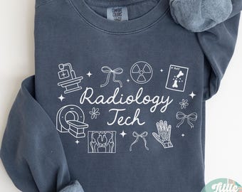 Puede incluir: Sudadera azul denim con ilustraciones gráficas blancas y texto "Radiology Tech". El diseño incluye equipos de imagen médica, una mano esquelética y lazos. La sudadera es de Comfort Colors.