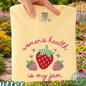 May include: A pale yellow t-shirt featuring a red strawberry graphic with green gingham leaves and two smaller strawberry accents. The text reads women's health is my jam in red script. The shirt has a Comfort Colors brand label at the neck.
