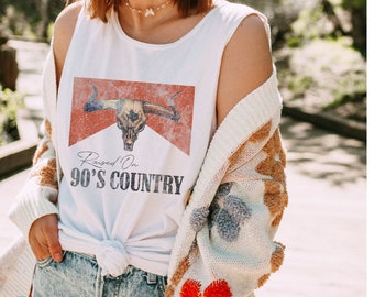 May include: White sleeveless tank top with a red, white, and blue graphic of a bull skull with the text "Raised On 90's Country".