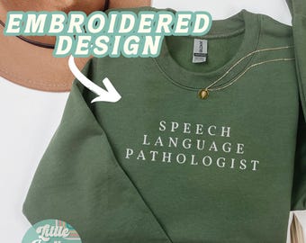 May include: Military green sweatshirt with the text "SPEECH LANGUAGE PATHOLOGIST" in white. The sweatshirt has a crew neck and long sleeves. A gold necklace with a pendant is draped on the sweatshirt. The words "EMBROIDERED DESIGN" are above the sweatshirt.
