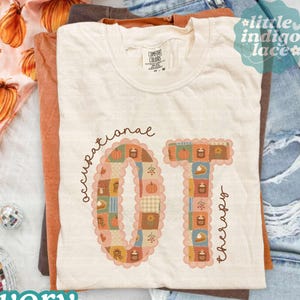May include: An ivory crew neck t-shirt featuring the text occupational therapy and the letters OT filled with a patchwork pattern of pumpkins, leaves, and autumn motifs. The shirt is layered over a brown shirt, a pumpkin print fabric, and blue denim jeans.