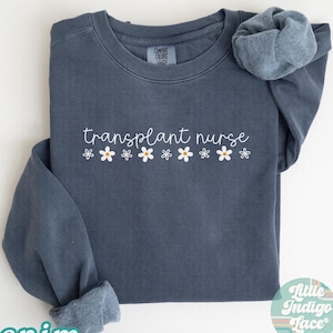 May include: A denim blue sweatshirt with the words "transplant nurse" in white script, accented by small white and yellow flower graphics. The sweatshirt has a crew neck and long sleeves, with the cuff of one sleeve rolled up. The word "denim" is printed in a teal color at the bottom left.