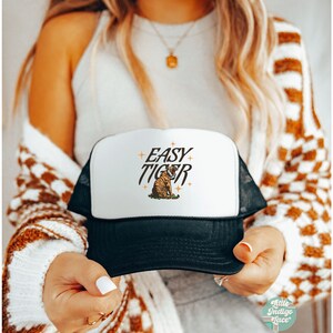 May include: A white and black trucker hat with a black mesh back. The front of the hat features a tiger illustration and the text "EASY TIGER" in a retro font.