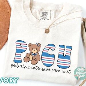 May include: Ivory t-shirt with a graphic of the letters "PCU" in blue, pink, and white stripes. A brown teddy bear with a pink bow is in the center of the letters. The text "pediatric intensive care unit" is below the graphic. The text "Little Indigo Face" is in the bottom right corner of the image.