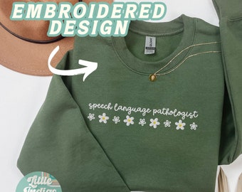May include: A military green sweatshirt with the text "speech language pathologist" embroidered in white with a row of white daisies below the text. The sweatshirt is laid flat on a white surface with a brown hat and a green plant in the background.