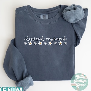 May include: A denim blue sweatshirt with the text "clinical research" in white lettering and a row of white daisies with yellow centers.