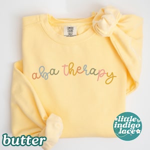May include: A butter-yellow sweatshirt with the words "aba therapy" in pastel colors. A matching scrunchie is on the shoulder. The word "butter" is at the bottom left, and a small logo is at the bottom right.