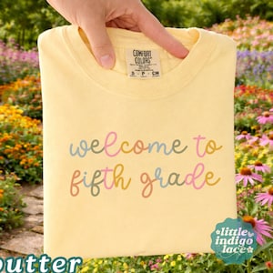 May include: A pale yellow t-shirt featuring the text "welcome to fifth grade" in a multi-colored cursive font. The shirt has a Comfort Colors brand label at the neck, indicating size small, and is made of 100% cotton material.