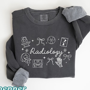 May include: A dark gray sweatshirt with the word "Radiology" in white script, surrounded by white medical-themed graphics. The sleeves have a gray, fuzzy lining. The word "pepper" is at the bottom left.