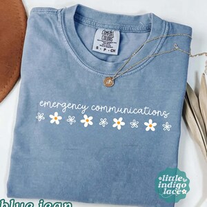 May include: A blue jean colored short-sleeve t-shirt with a crew neckline. The shirt features the white text "emergency communications" printed above a row of white and yellow daisy flower icons. A gold-toned necklace rests on the fabric.