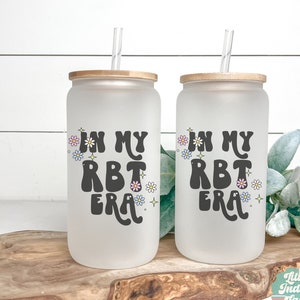 May include: Two frosted glass tumblers with bamboo lids and straws. The tumblers have the text "In My RBT Era" printed on them with a daisy design.