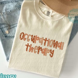 May include: An ivory crew neck t-shirt with the text "occupational therapy" printed in a burnt orange color. The letters feature a mix of polka dot, plaid, and striped patterns. The shirt has a Comfort Colors brand label at the inner neckline.
