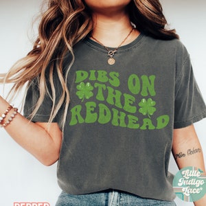 Op de afbeelding: Een donkergrijs T-shirt met een groene afbeelding waarop "DIBS ON THE REDHEAD" staat met klavertjesvier designs.