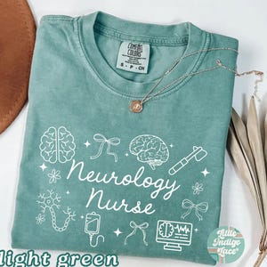 May include: Light green t-shirt with the words "Neurology Nurse" and related medical graphics in white. The shirt has a crew neck and short sleeves. A silver necklace with a pendant is draped across the shirt.
