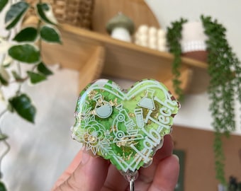 May include: Green and gold heart-shaped resin badge holder with a floral design and the word "Lab Tech" written on it. The badge holder is attached to a clear plastic retractable badge reel.