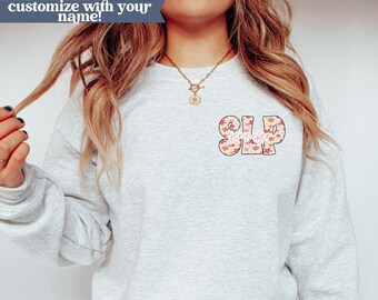May include: A light grey sweatshirt with a floral patterned design that says "SLP" with the name "Jennifer" written underneath. The sweatshirt is made by Ash-Gildan and is size 1800.