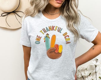 May include: Pale grey t-shirt featuring the text "ONE THANKFUL BCBA" in a circular arrangement. The graphic shows a hand making a peace sign, with a turkey design on the index finger. The shirt is made of a soft material.
