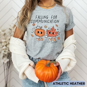 May include: Athletic heather gray t-shirt with the text "FALLING FOR COMMUNICATION" and a cartoon pumpkin and leaf holding hands. The shirt also has the text "SLP EDITION". An orange pumpkin is held in the hands.